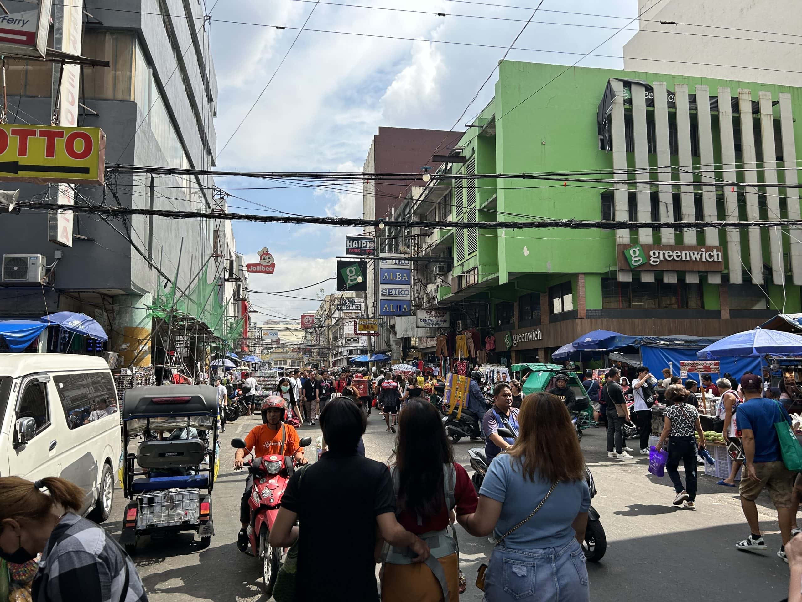 Busy street scene in Manila, Philippines, with crowds of pedestrians, market stalls, motorcycles, and shops lining the road under tangled overhead power lines.