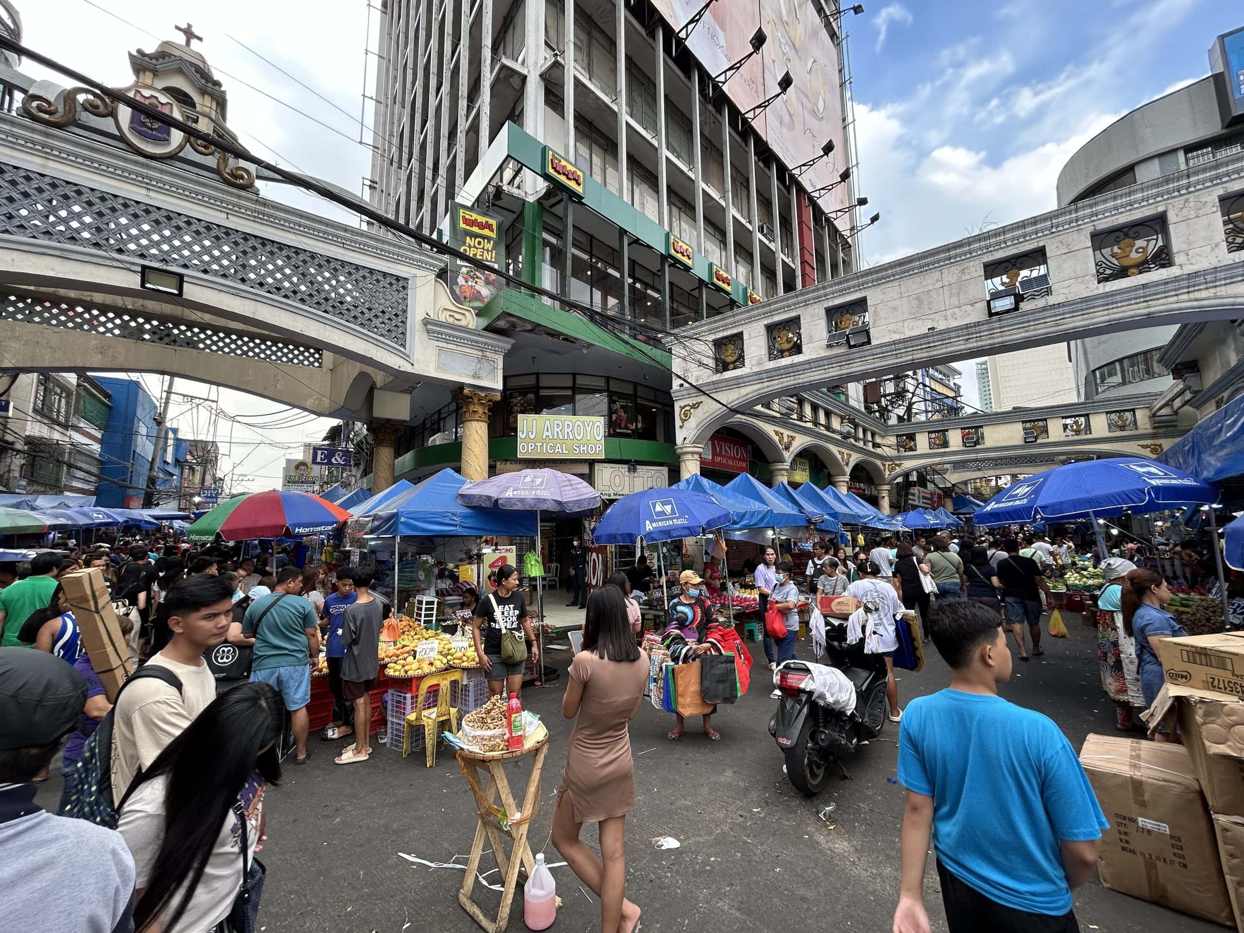 Crowded street market in Manila, Philippines, with fruit vendors, blue umbrellas, shoppers, and busy city buildings in the background.