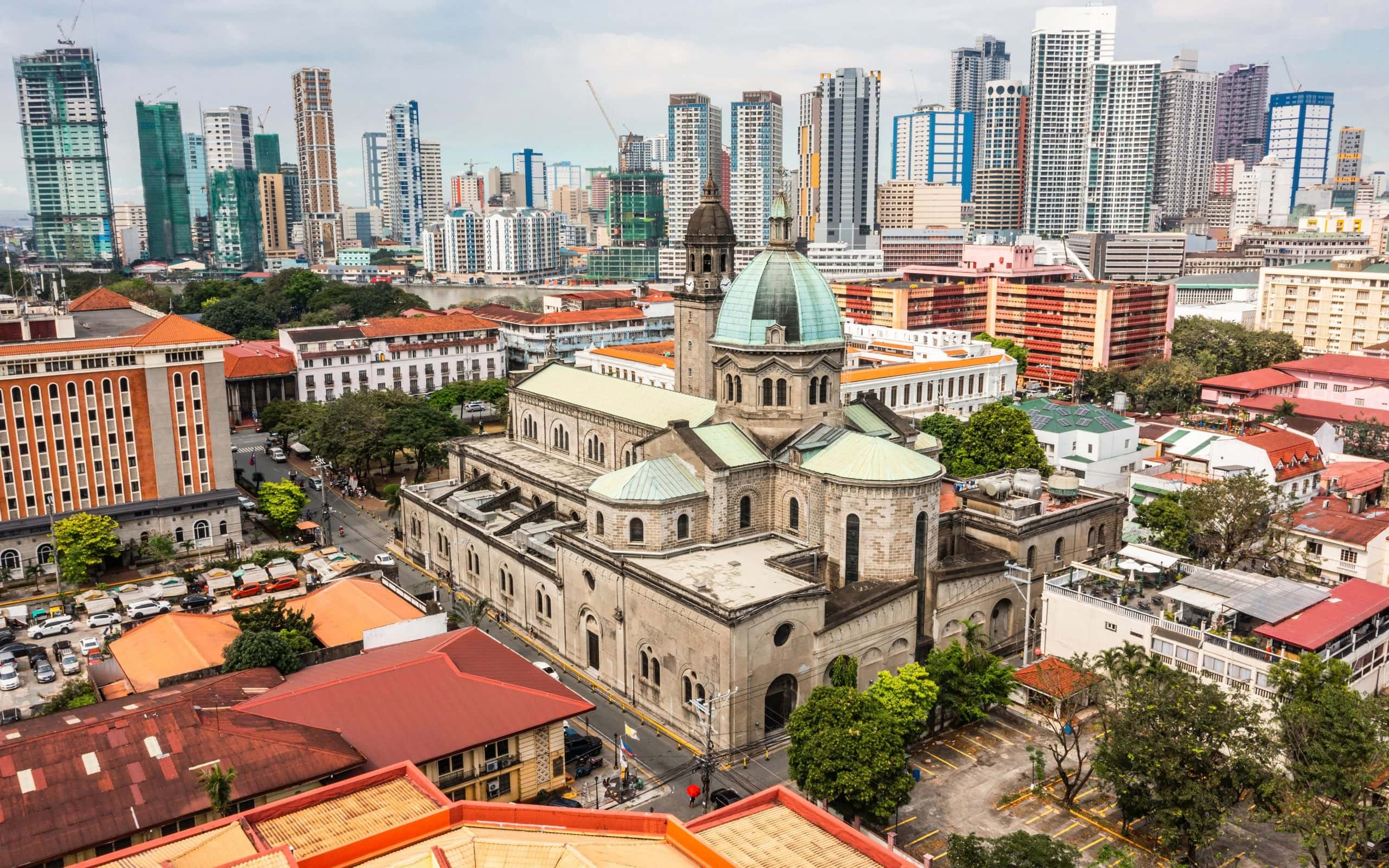 Aerial view of Manila Cathedral in Intramuros with the modern Manila skyline in the background, showing historic architecture surrounded by skyscrapers.