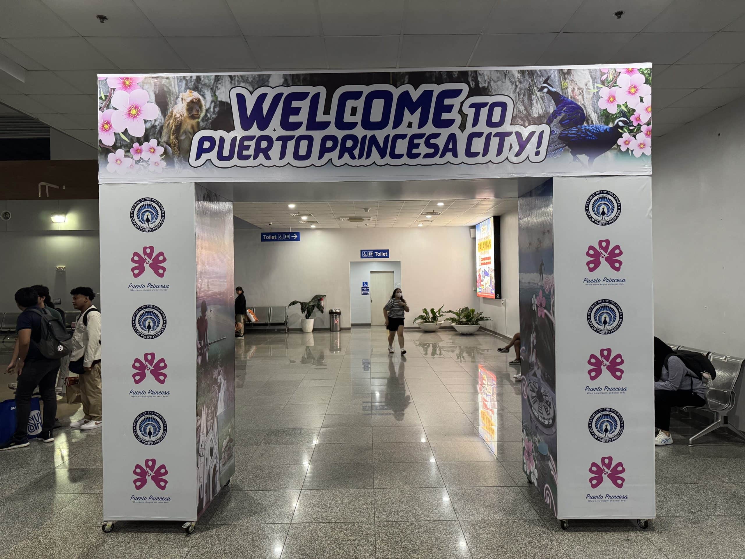 Welcome arch inside Puerto Princesa Airport reading “Welcome to Puerto Princesa City,” with travelers walking through the terminal and tropical graphics highlighting Palawan’s main gateway city.