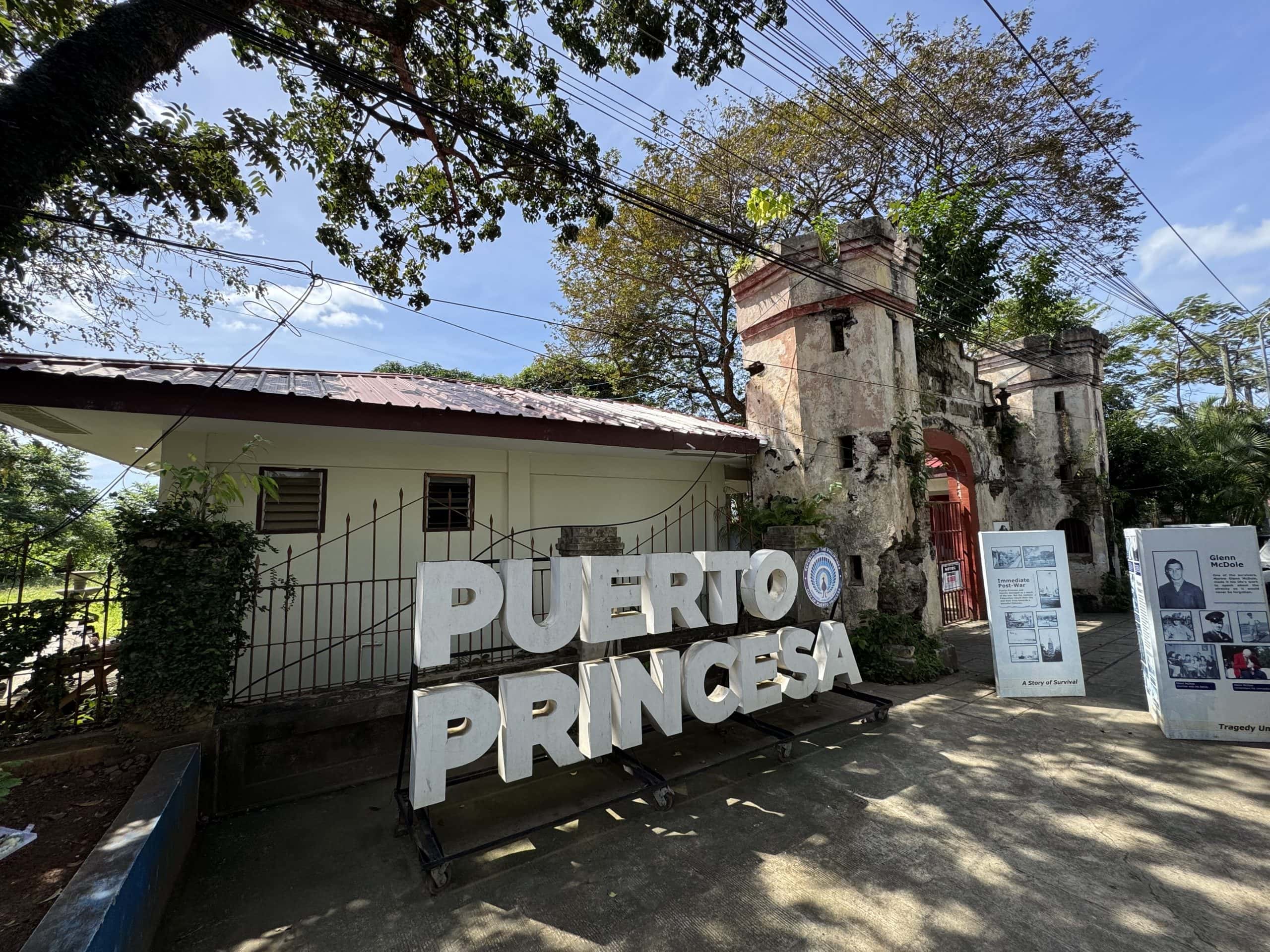 Historic entrance area in Puerto Princesa featuring old stone gate ruins, museum displays, and large Puerto Princesa sign surrounded by tropical trees in Palawan, Philippines
