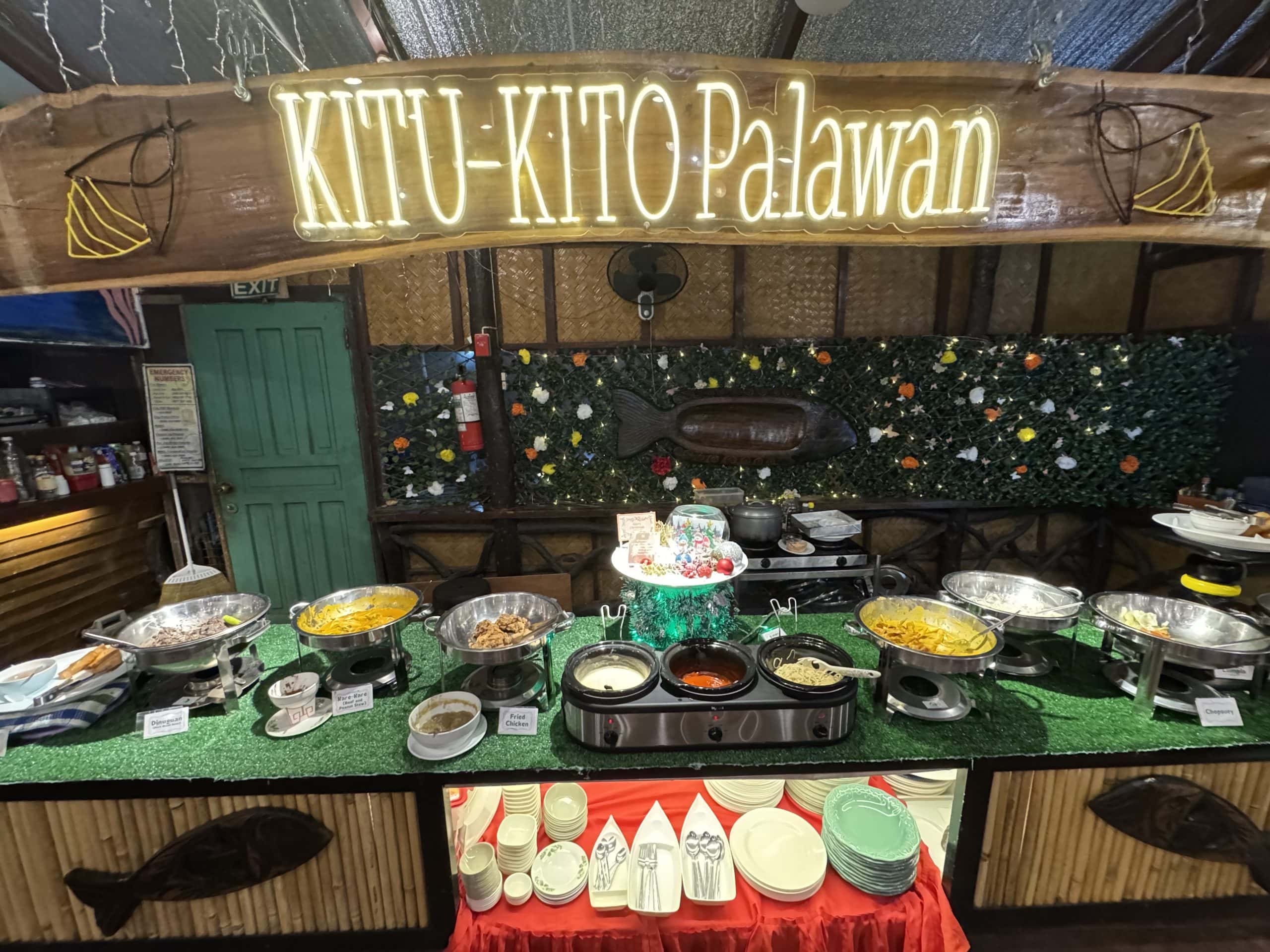 Buffet spread at Kitu Kito Palawan featuring traditional Filipino dishes under a glowing wooden sign inside a cozy tropical style restaurant in Puerto Princesa, Palawan