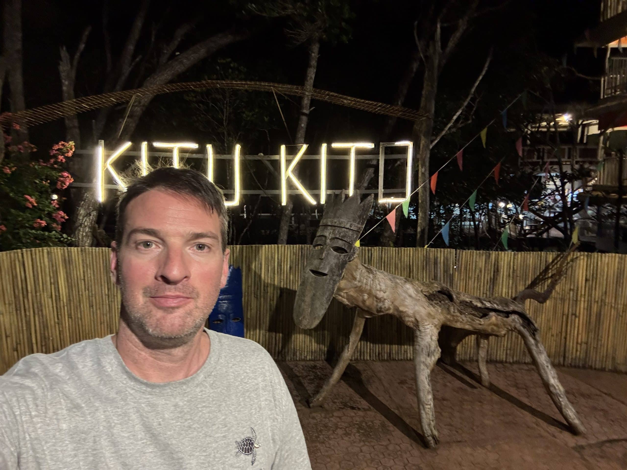 Traveler posing at night in front of the glowing Kith Kito sign with wooden tribal art sculptures and tropical surroundings in Puerto Princesa, Palawan, Philippines