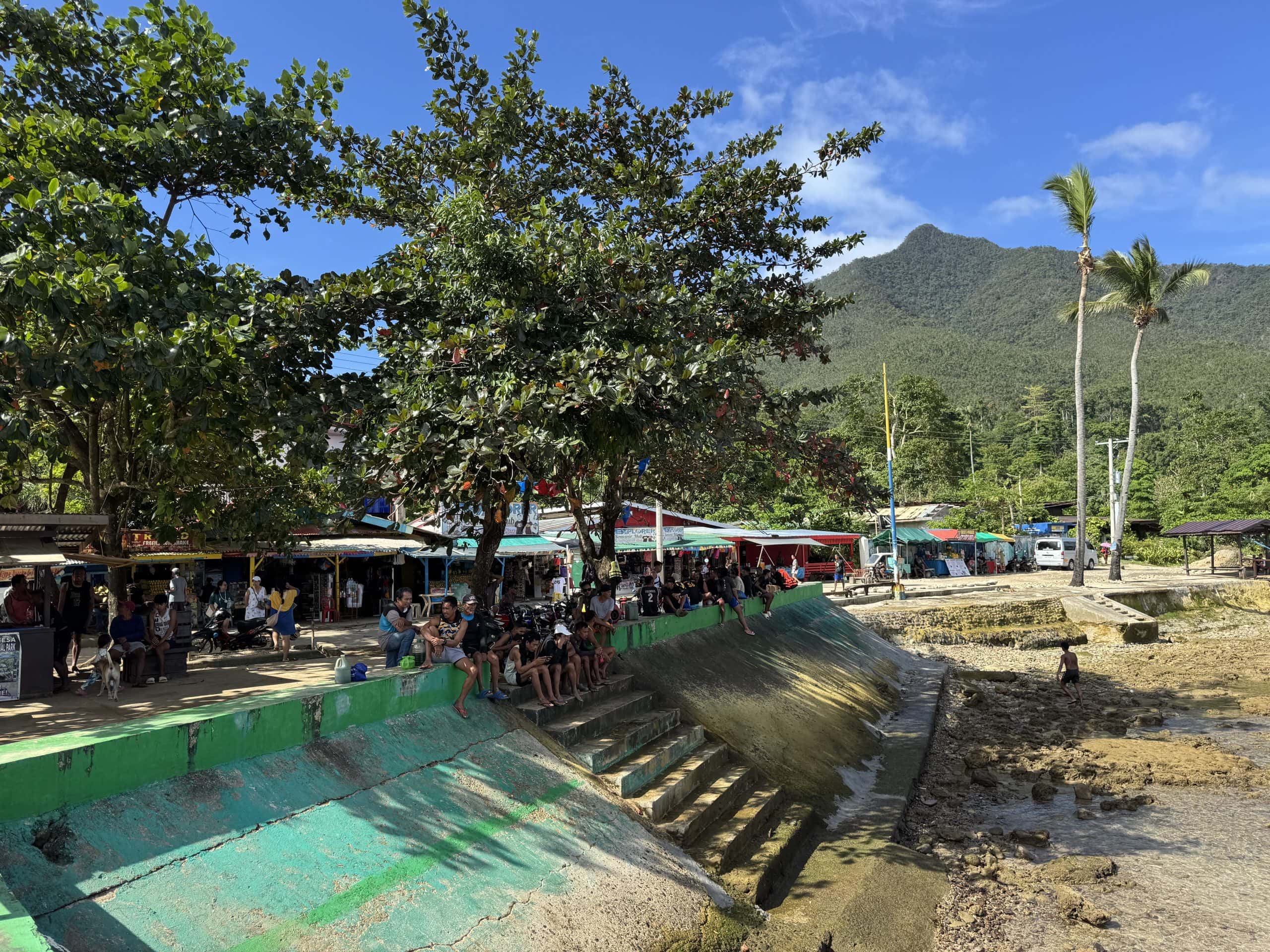 Local villagers and visitors relaxing along the waterfront near the Puerto Princesa Underground River with market stalls, palm trees, and jungle mountains in Palawan, Philippines