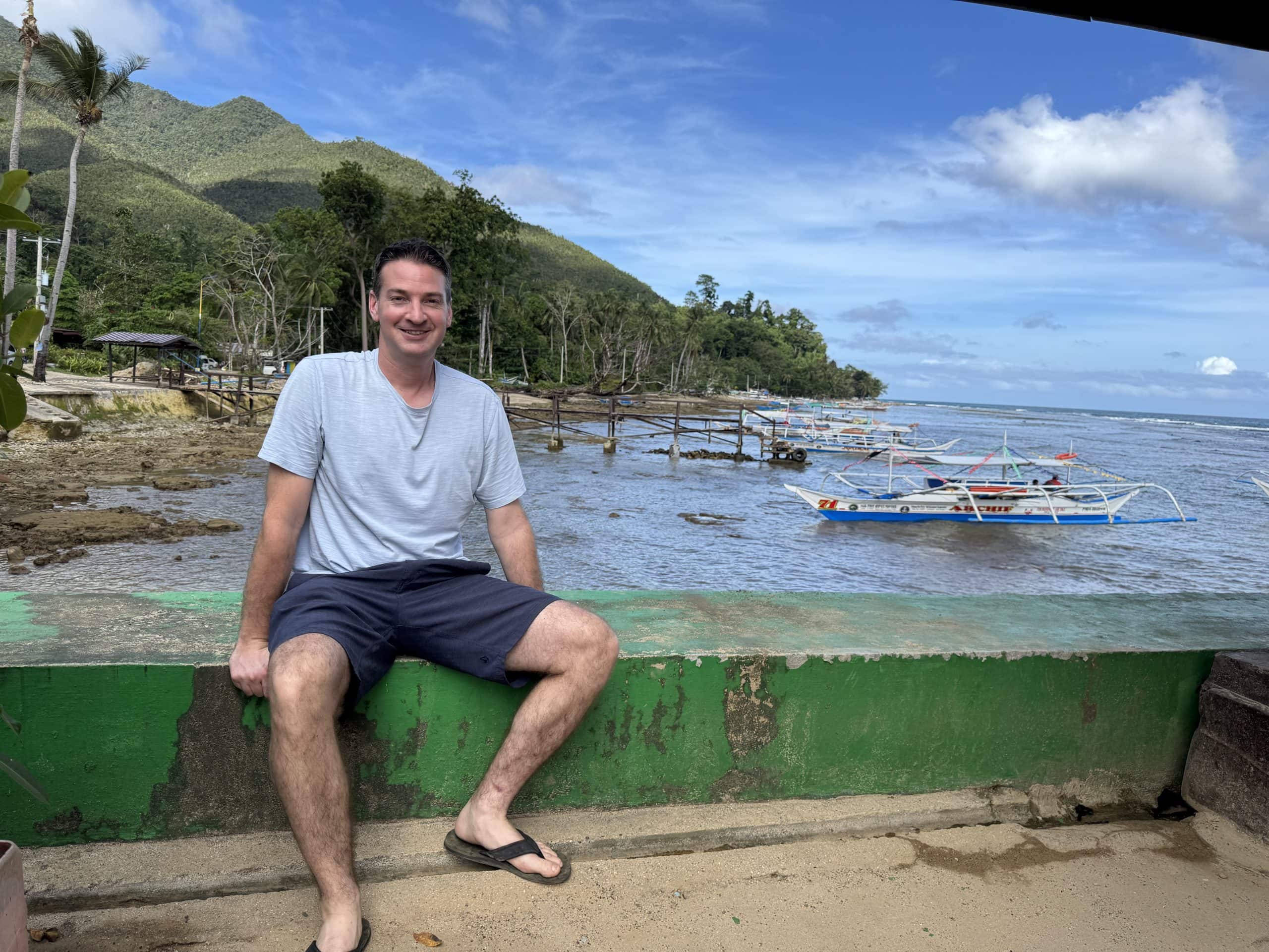 Traveler sitting along a coastal seawall in Palawan with traditional Filipino outrigger boats anchored in shallow water, a wooden pier stretching into the bay, and lush green mountains rising behind the shoreline.