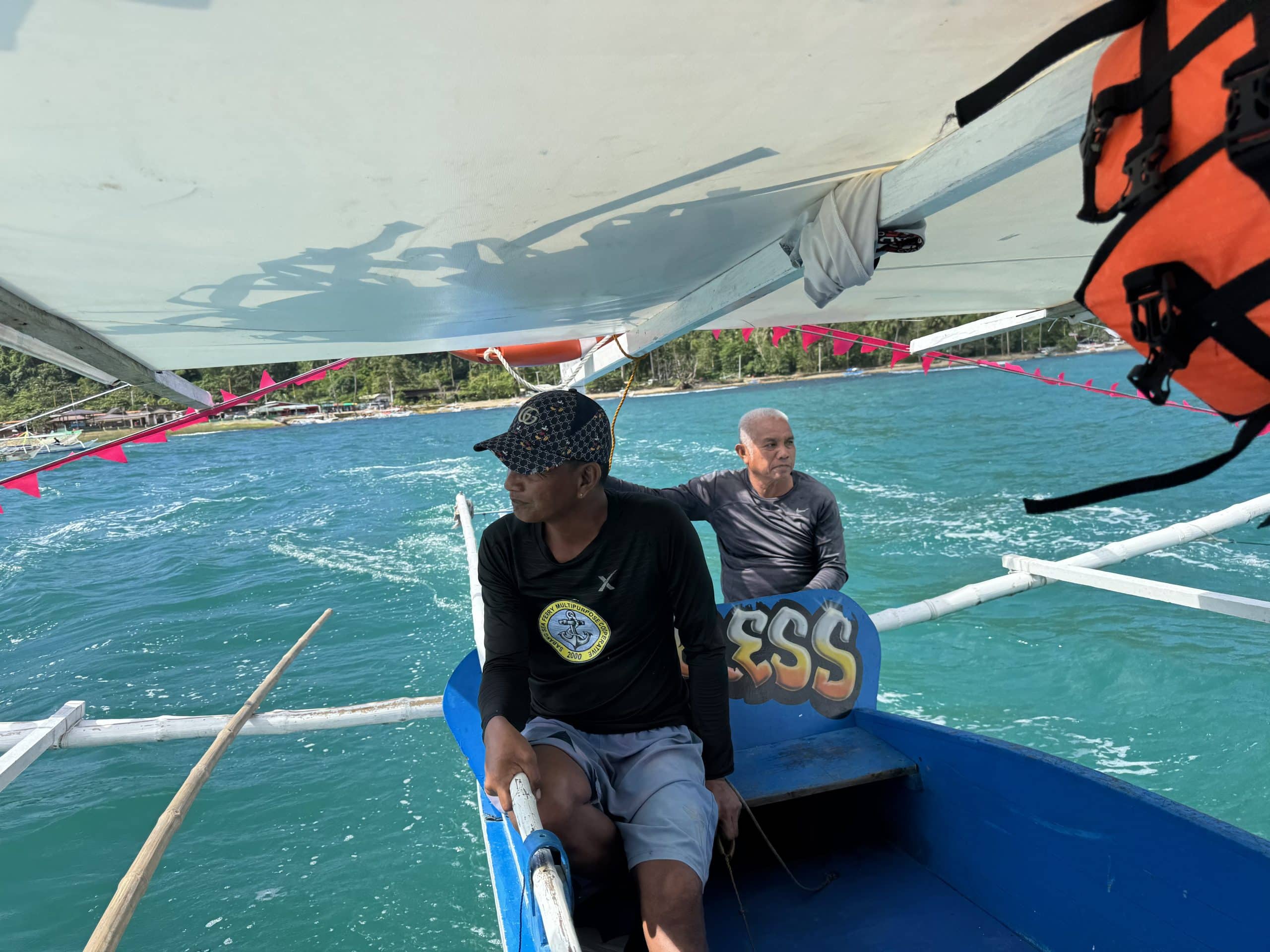 Boat crew guiding a traditional outrigger across turquoise waters toward the Puerto Princesa Underground River with tropical shoreline in the background in Palawan, Philippines