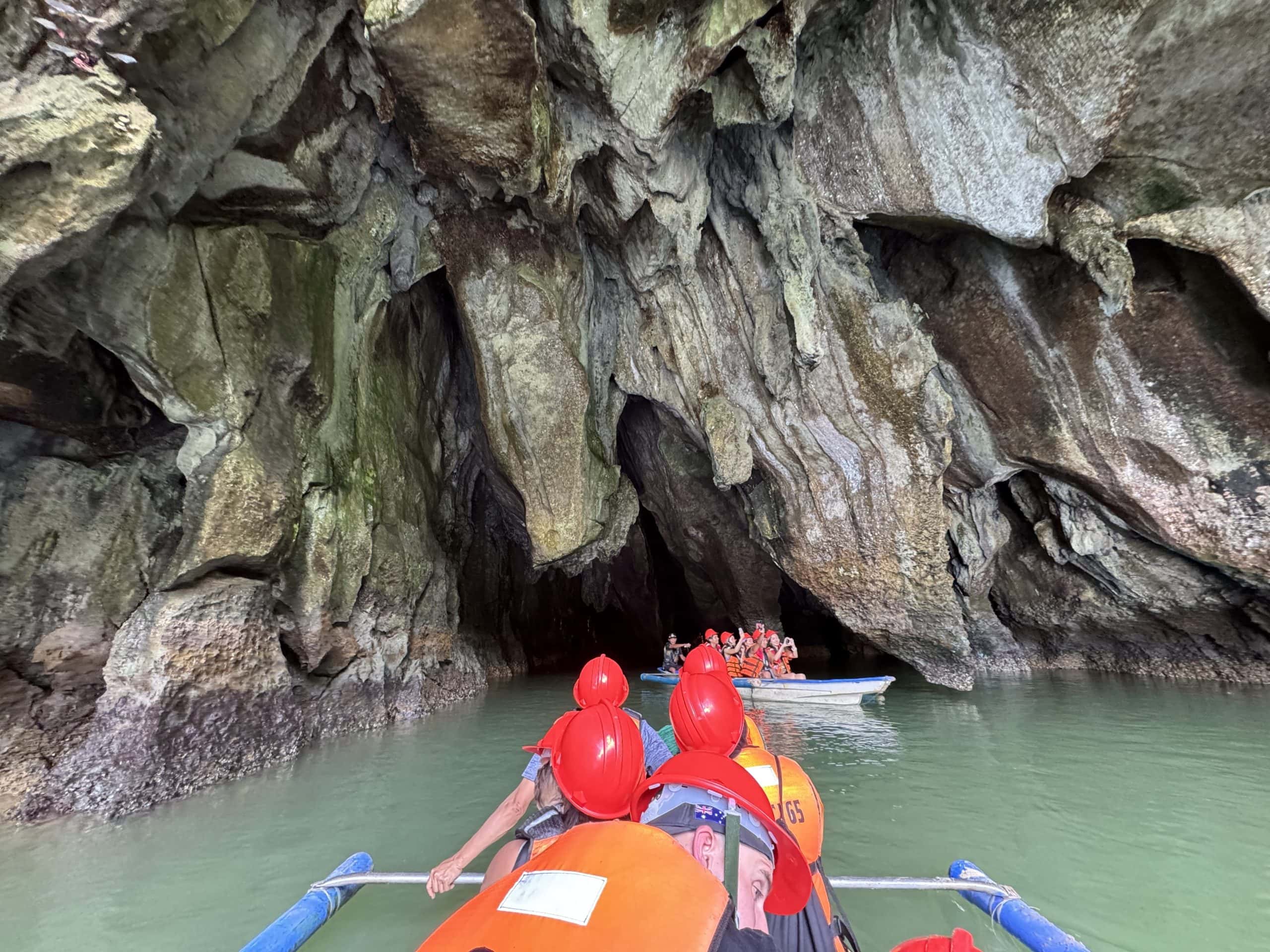 Tourists wearing helmets and life jackets paddling into the entrance of an underground river cave in Palawan, with towering limestone rock formations rising above calm green water inside the cavern.