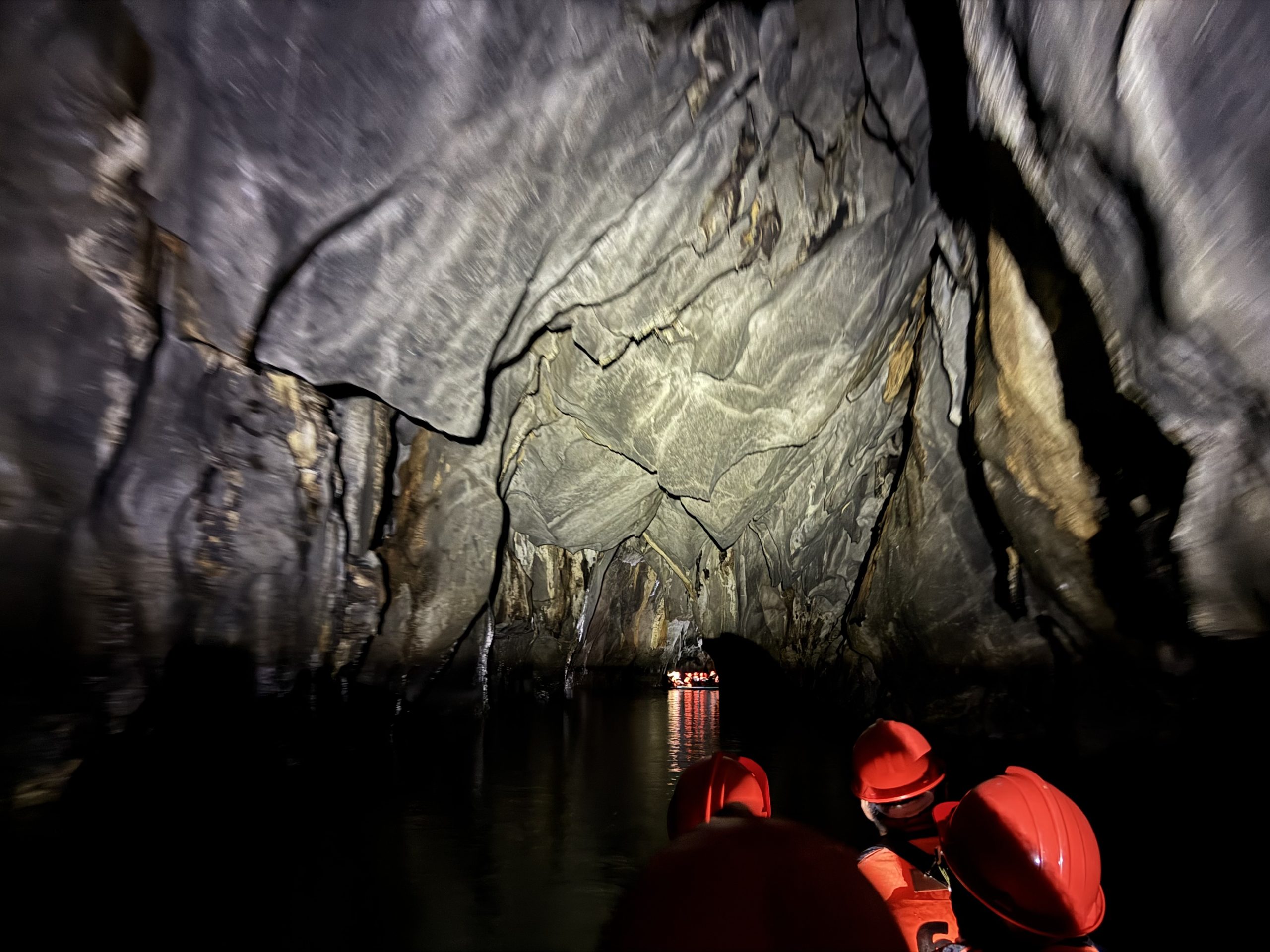Boat gliding through the dark interior of the Puerto Princesa Underground River cave, with dramatic limestone ceilings illuminated by tour lights and visitors wearing helmets and life jackets reflected on the calm water.