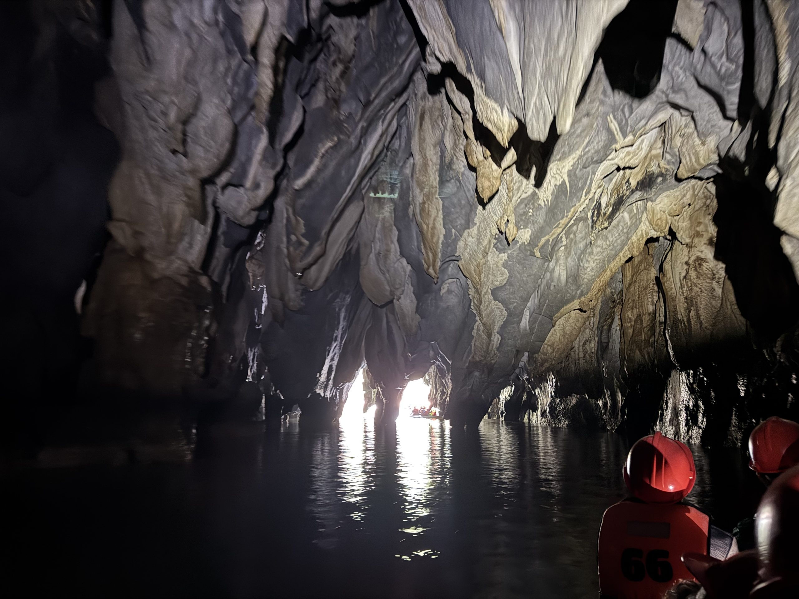 Inside the Puerto Princesa Underground River showing towering limestone cave walls, reflective water, and visitors in helmets drifting toward the lighted cave opening in Palawan, Philippines