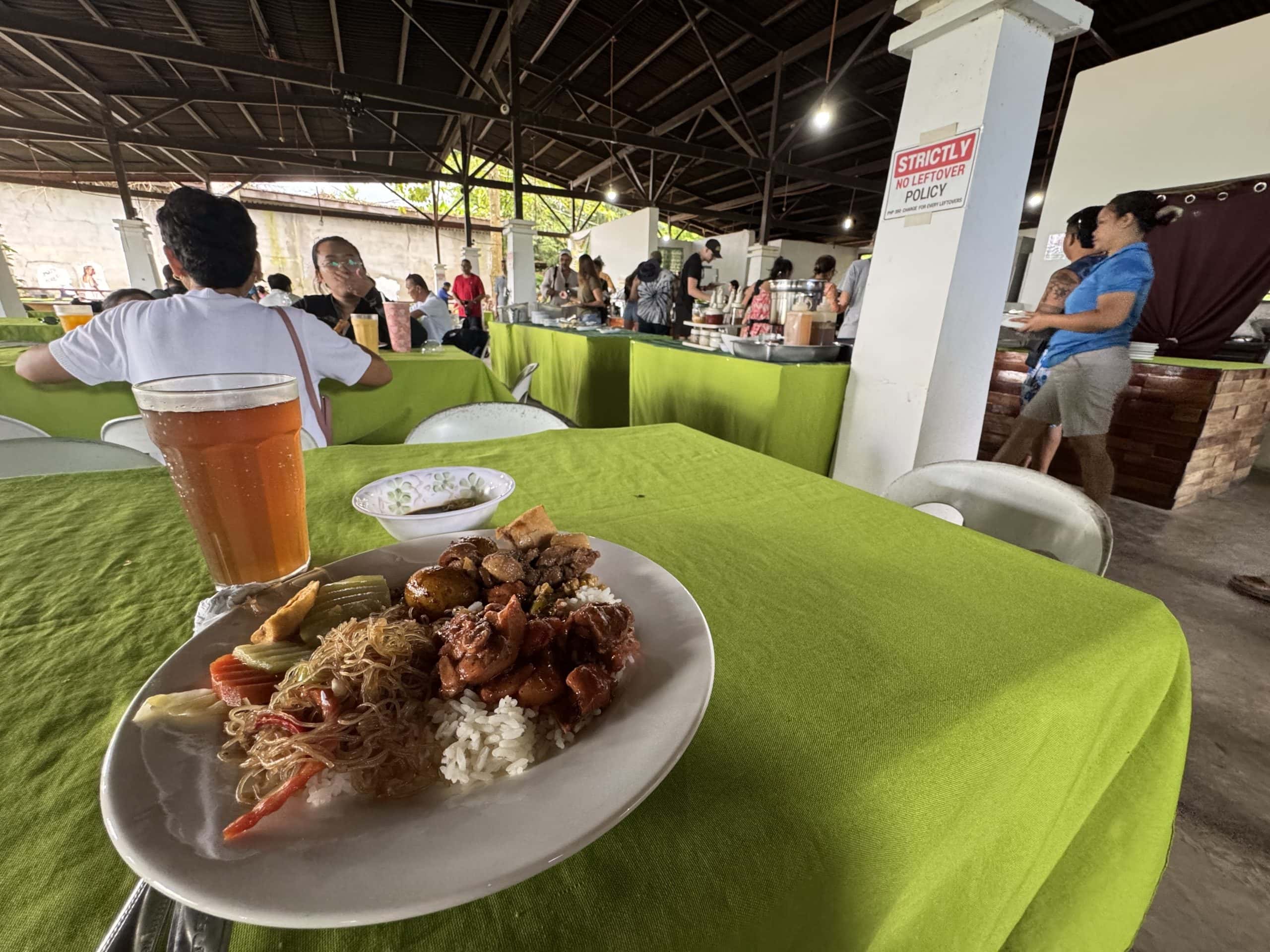 Buffet lunch at a local restaurant near the Puerto Princesa Underground River featuring Filipino dishes, rice, noodles, and a cold drink with travelers dining in the background in Palawan, Philippines
