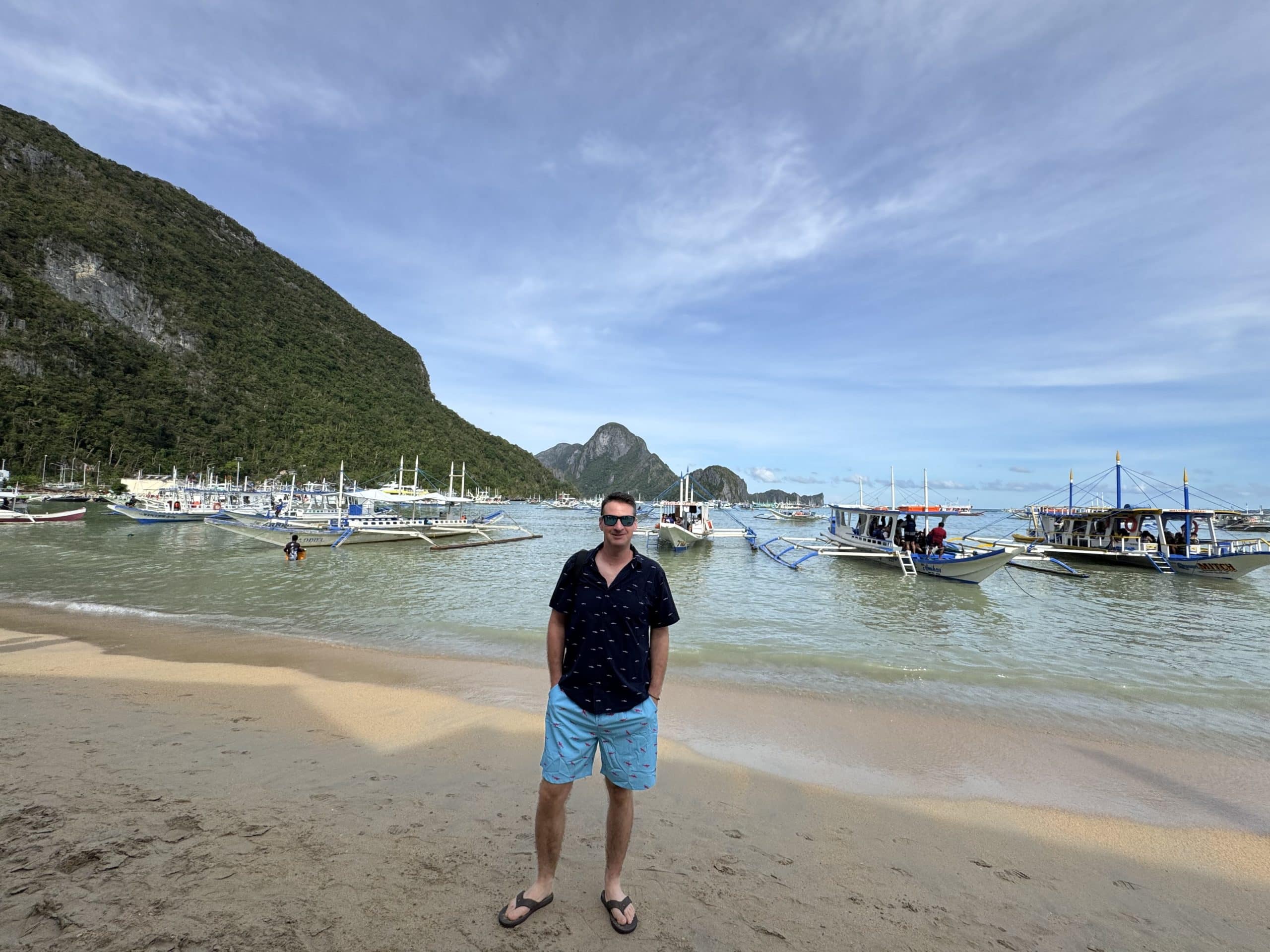 Tropical beach scene in El Nido, Palawan with crystal clear water, traditional Filipino outrigger boats anchored along the shore, limestone cliffs rising in the background, and a traveler standing barefoot on the sand under a bright blue sky.