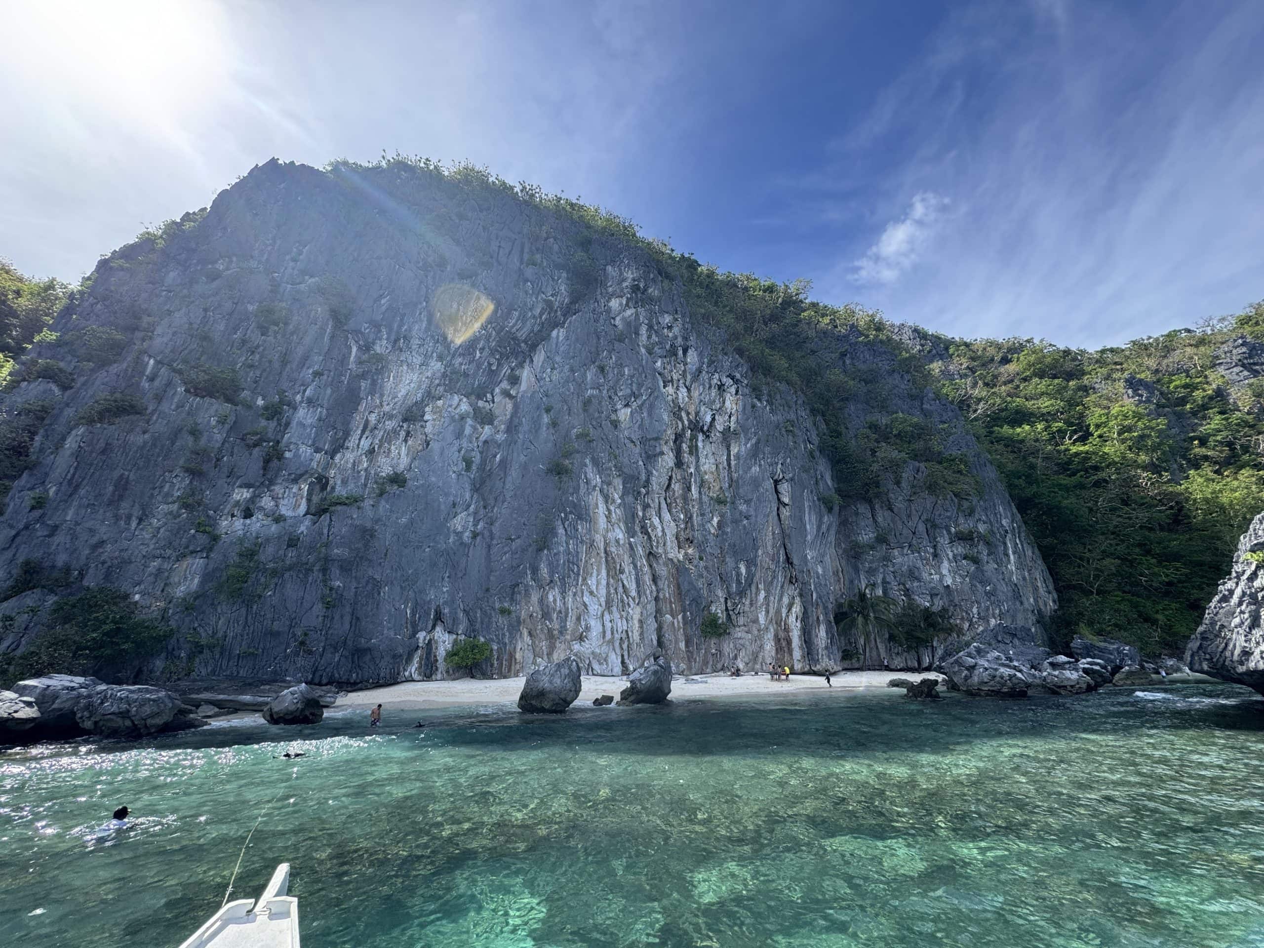 Massive limestone cliff towering over a secluded white sand beach in El Nido, Palawan, with crystal clear turquoise water in the foreground and lush greenery clinging to the rocky walls under a bright tropical sky.