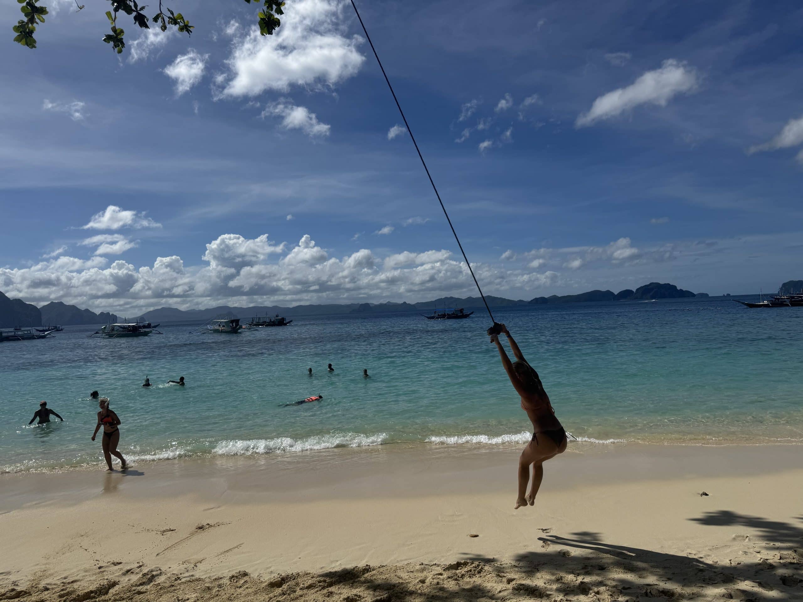 Traveler swinging on a rope over soft white sand into crystal clear turquoise water in El Nido, Palawan, with island hopping boats anchored offshore and limestone islands lining the horizon under a bright blue sky.