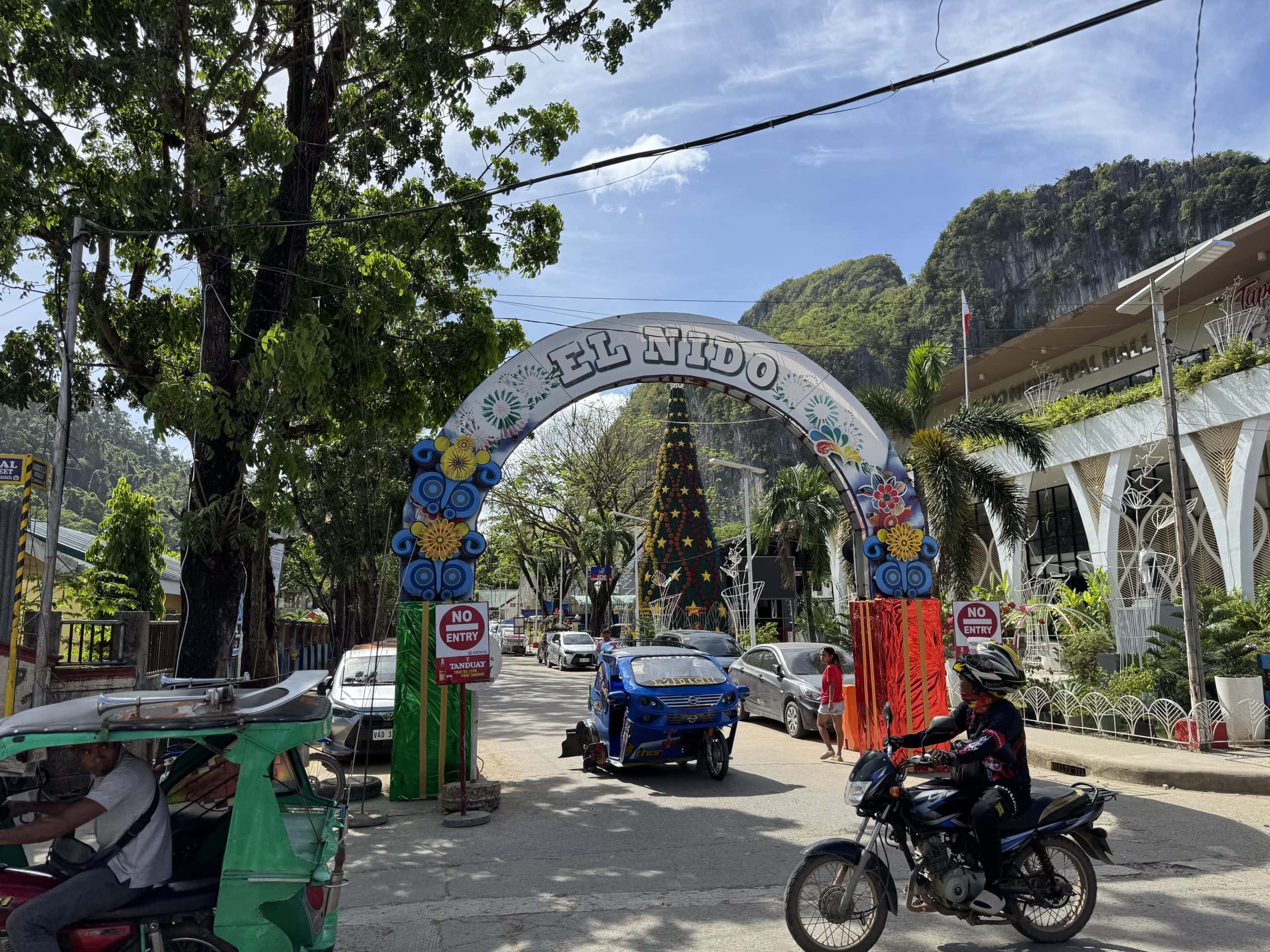 Colorful welcome arch reading “El Nido” at the entrance to town, with tricycles and motorbikes passing beneath it, limestone cliffs rising in the background, and palm trees lining the busy street in Palawan.