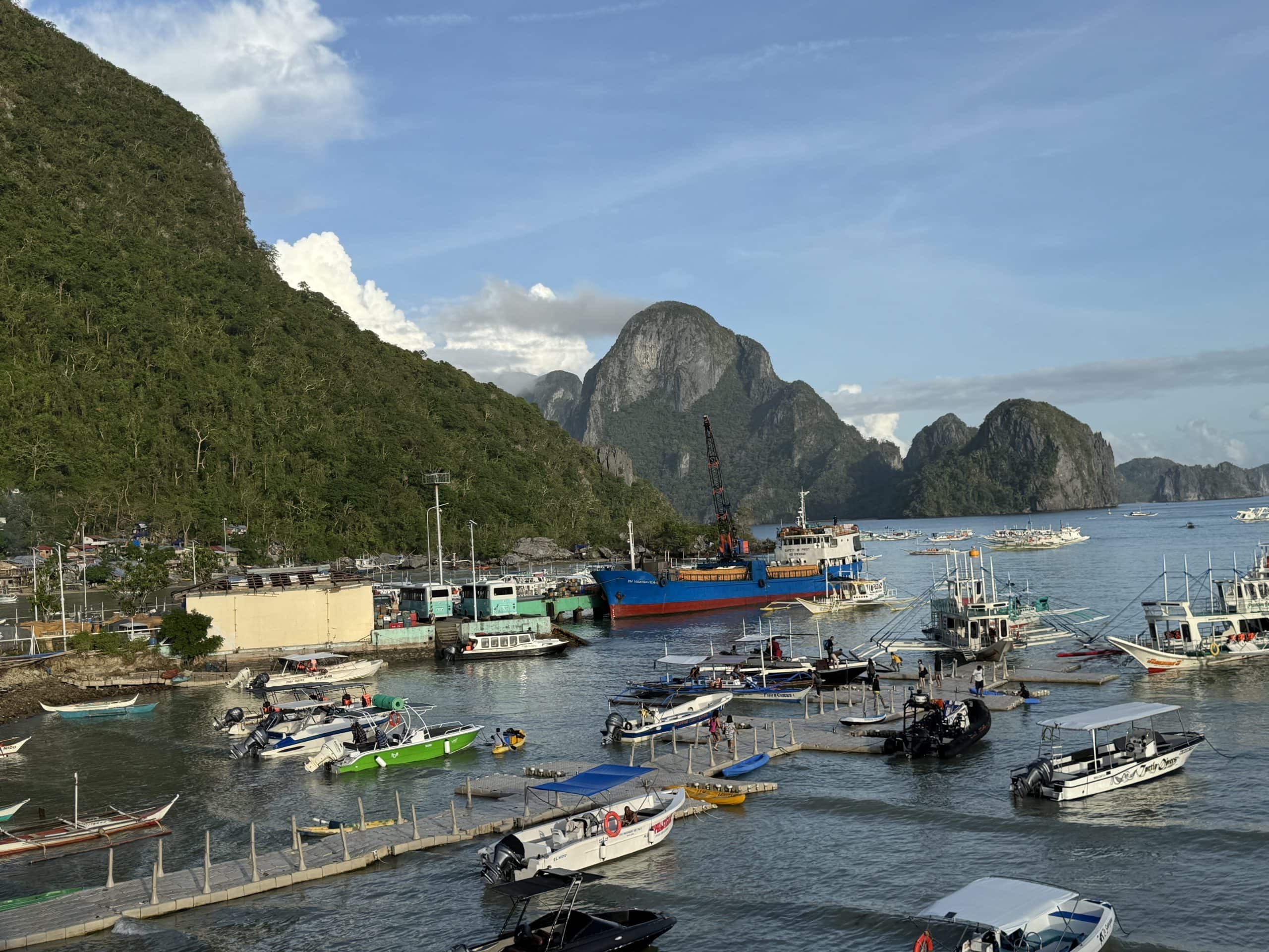 Busy harbor in El Nido, Palawan filled with tour boats and small docks, backed by dramatic limestone mountains and lush green hills overlooking the turquoise bay under a bright tropical sky.