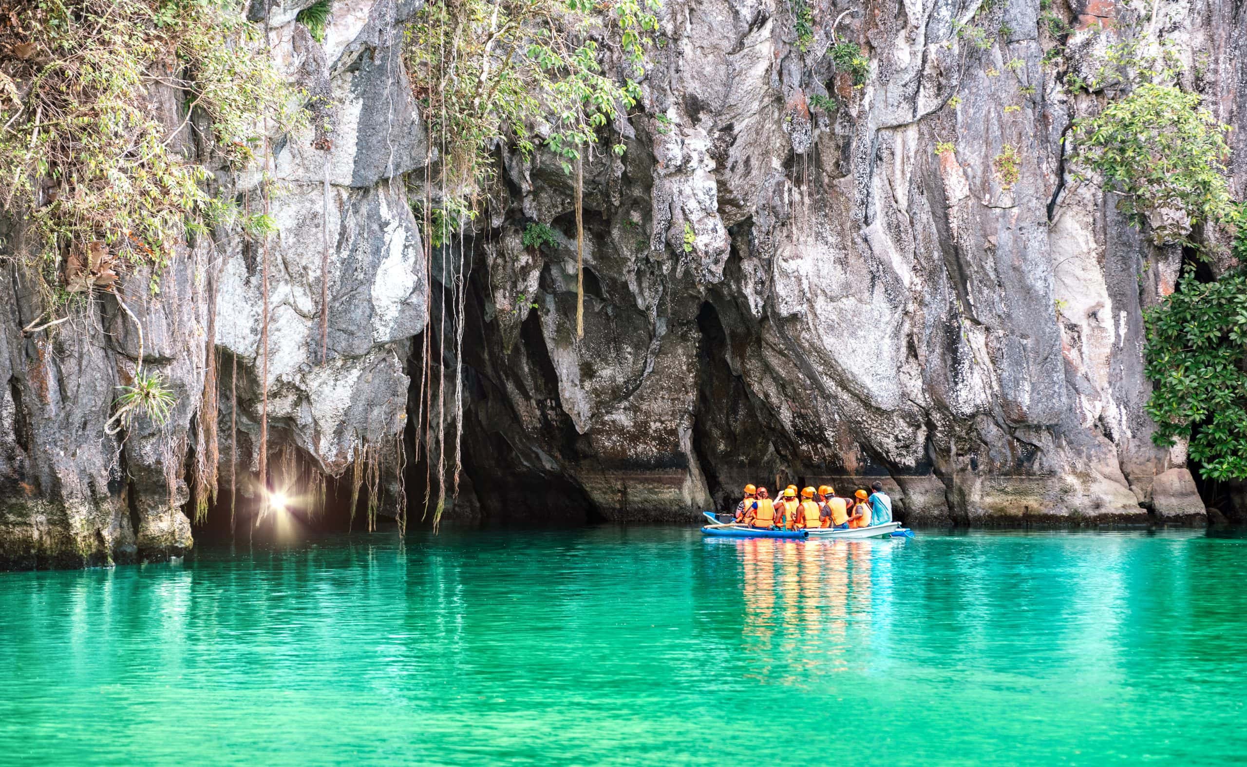 Small boat carrying visitors in life vests approaching the limestone cave entrance of the Puerto Princesa Underground River over clear turquoise water in Palawan, Philippines