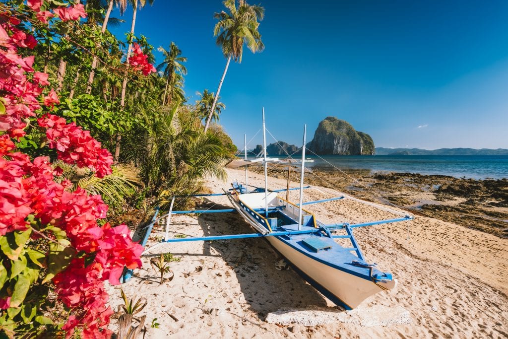 El Nido, Palawan, Philippines. Native banca boat and vibrant flowers at Las cabanas beach with amazing Pinagbuyutan island in background.
