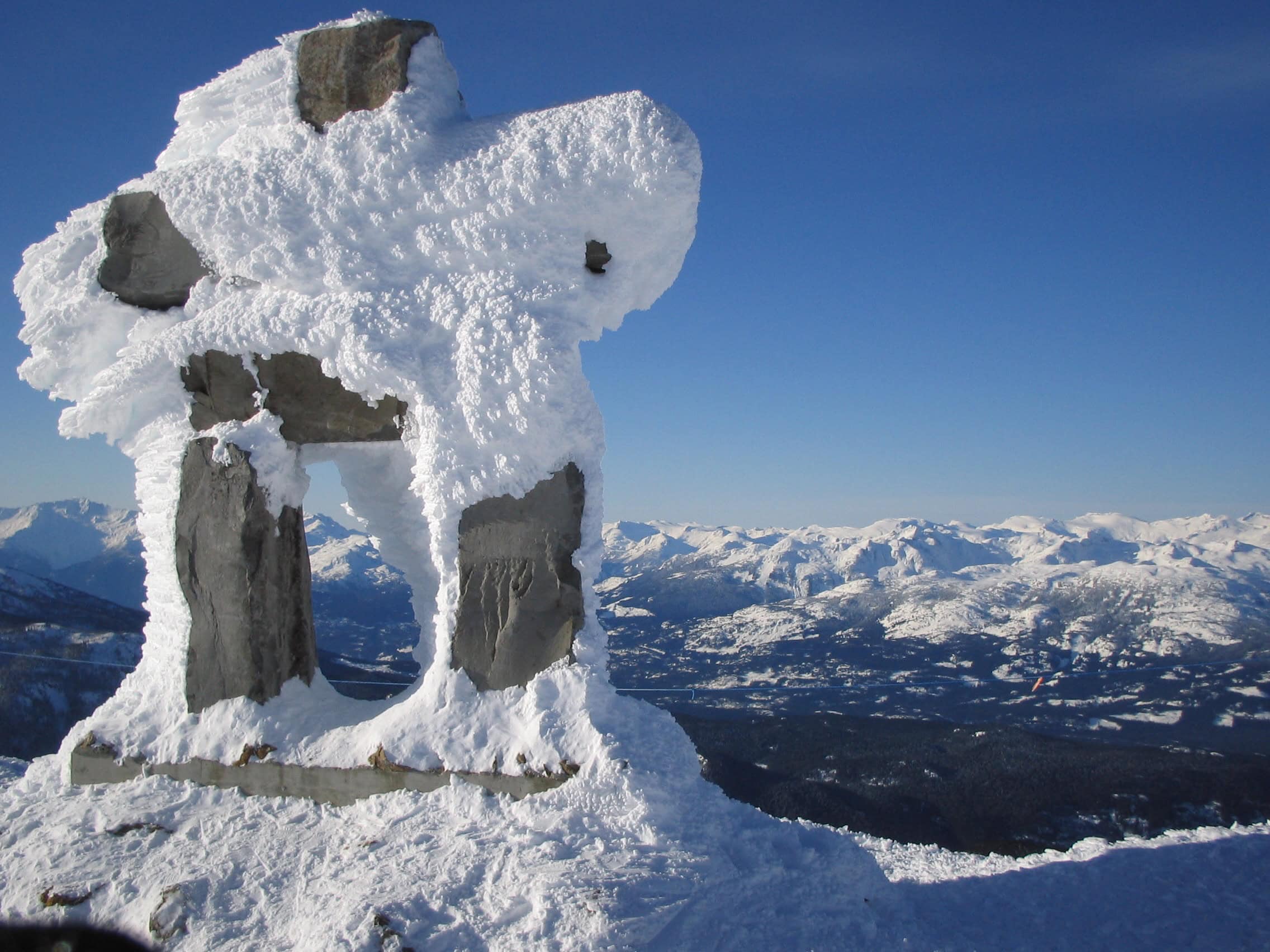 Snow covered Inukshuk at the summit of Whistler Mountain with panoramic views of the Coast Mountains and snowy alpine landscape under a clear blue sky