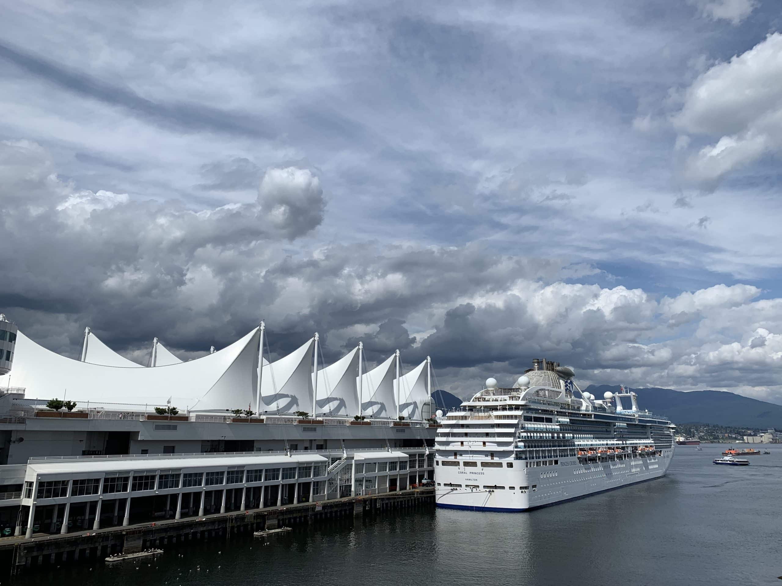 Cruise ship docked at Canada Place in Vancouver with the iconic white sail roof and dramatic coastal mountains under a cloudy sky