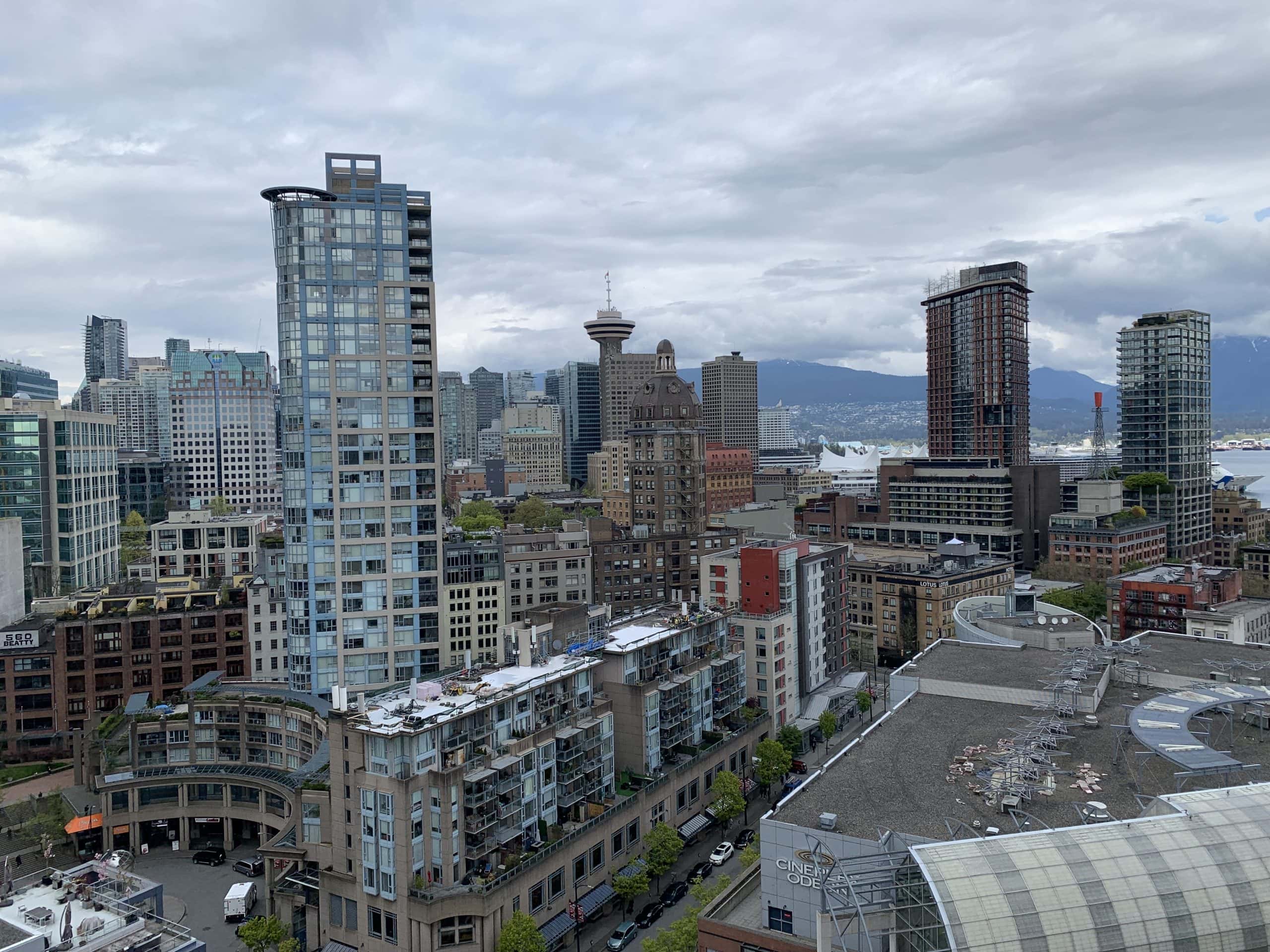 View of downtown Vancouver skyline with modern high rise buildings, Harbour Centre observation tower, and coastal mountains in the background under a cloudy Pacific Northwest sky.