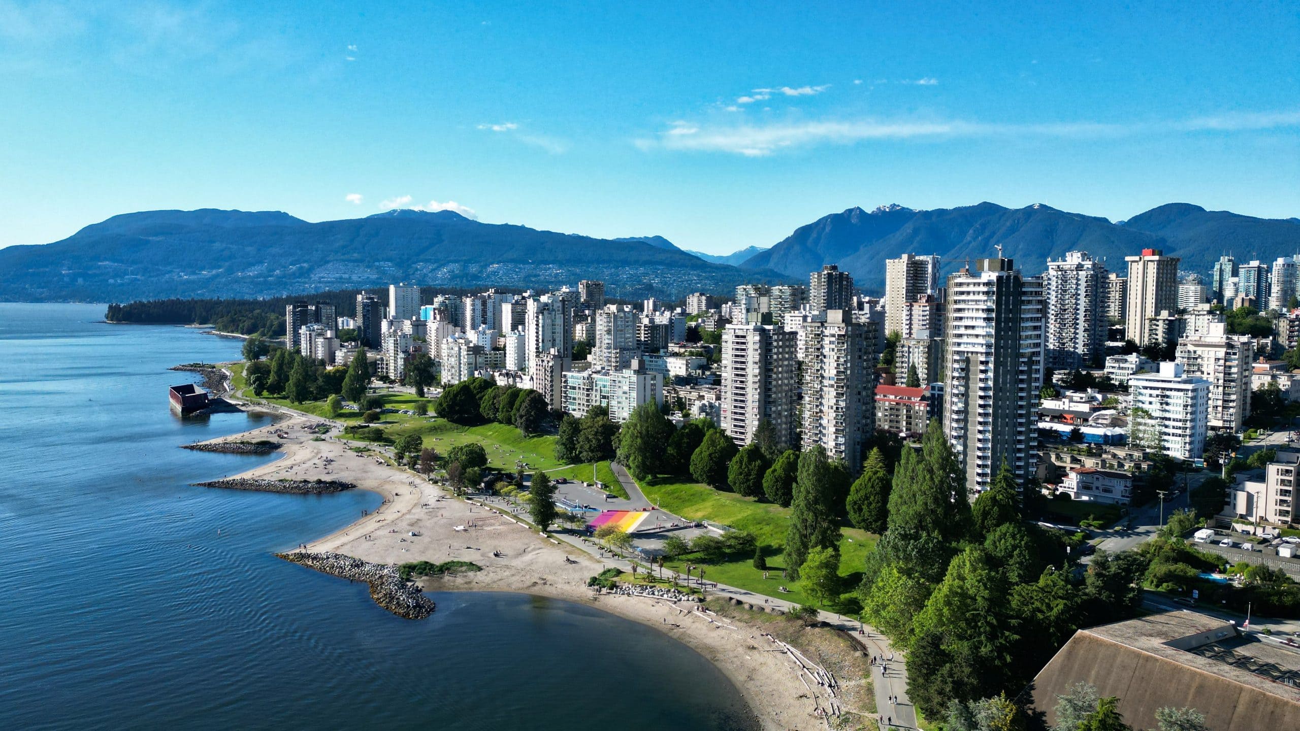 Aerial view of Vancouver’s West End with high rise residential buildings, English Bay beach, Stanley Park shoreline, and the North Shore Mountains under a clear blue sky.