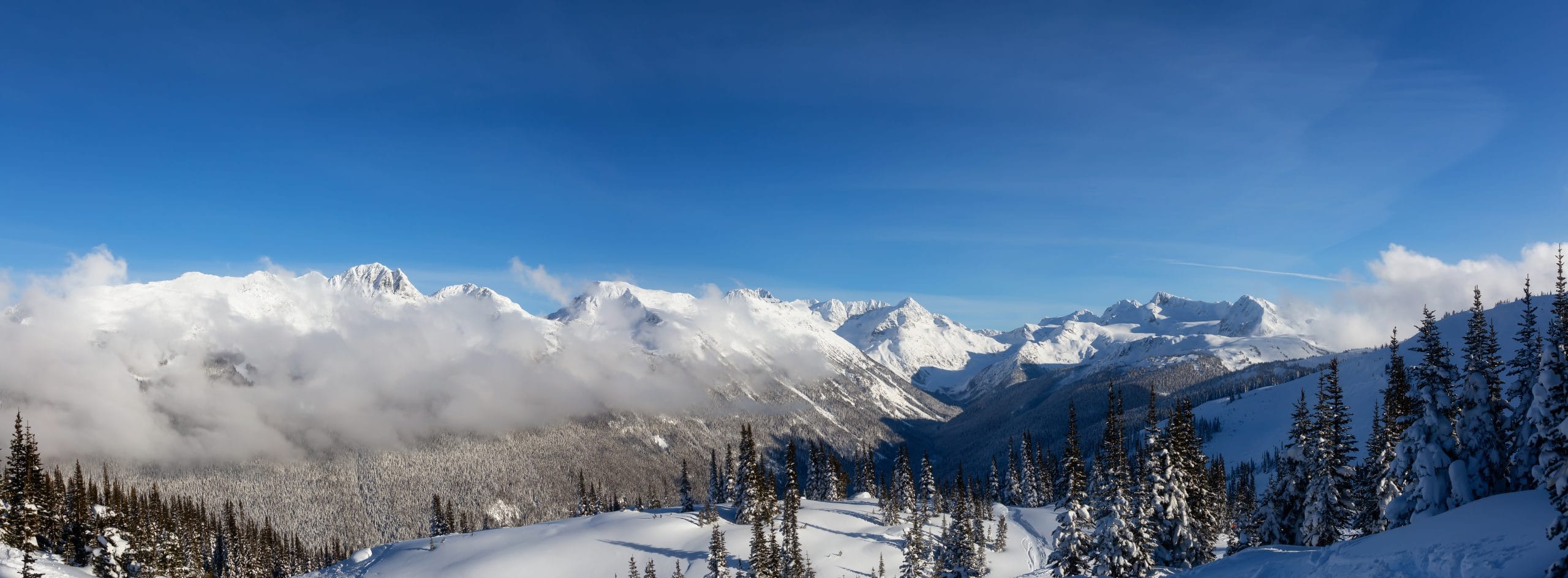 Panoramic view of snow covered mountains and alpine forest in Whistler, British Columbia with dramatic clouds rolling through the Coast Mountains under a clear blue sky