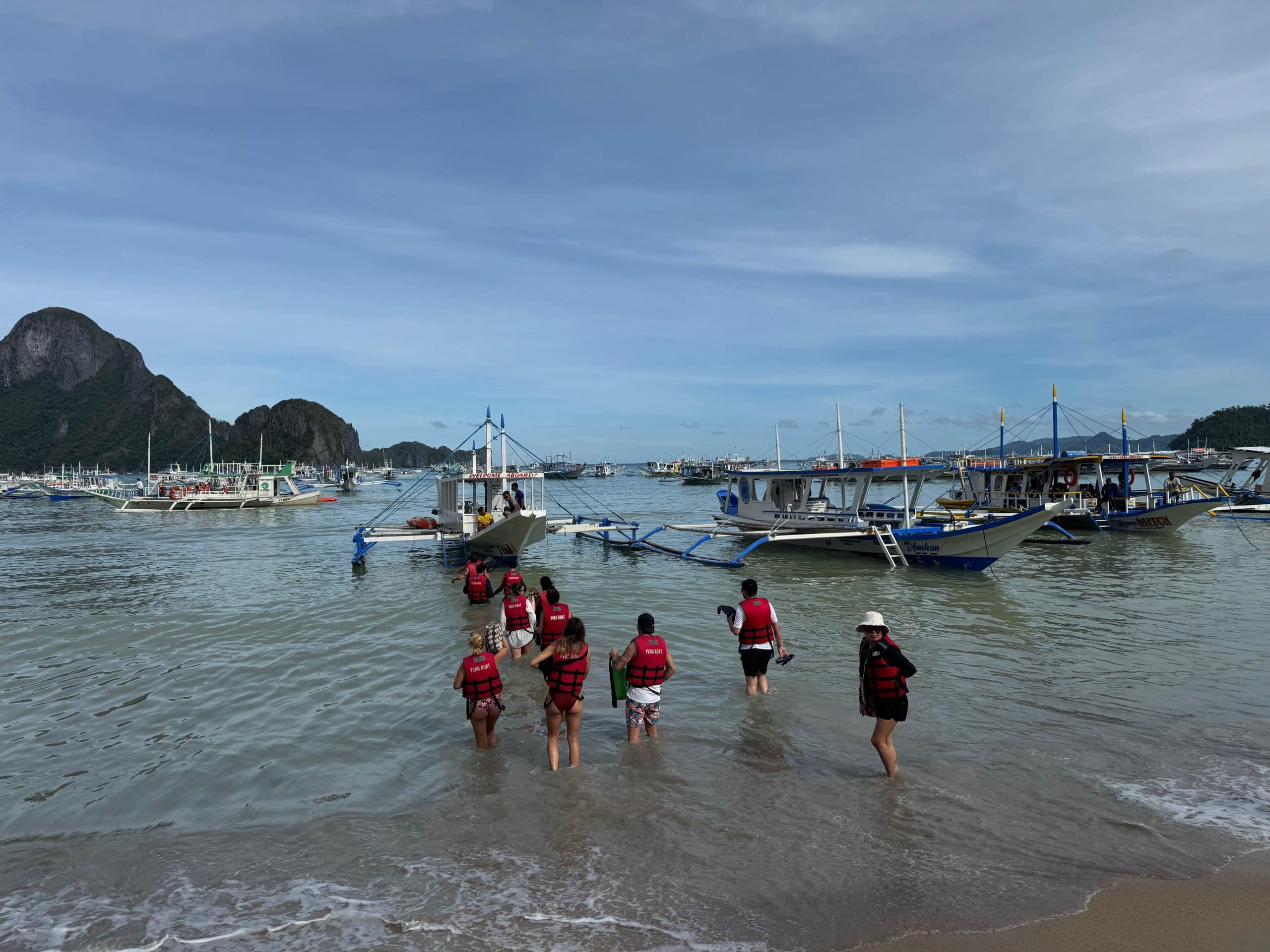 Tourists walking into the water to board traditional island hopping boats in El Nido with limestone mountains and anchored bangkas in the background in Palawan Philippines