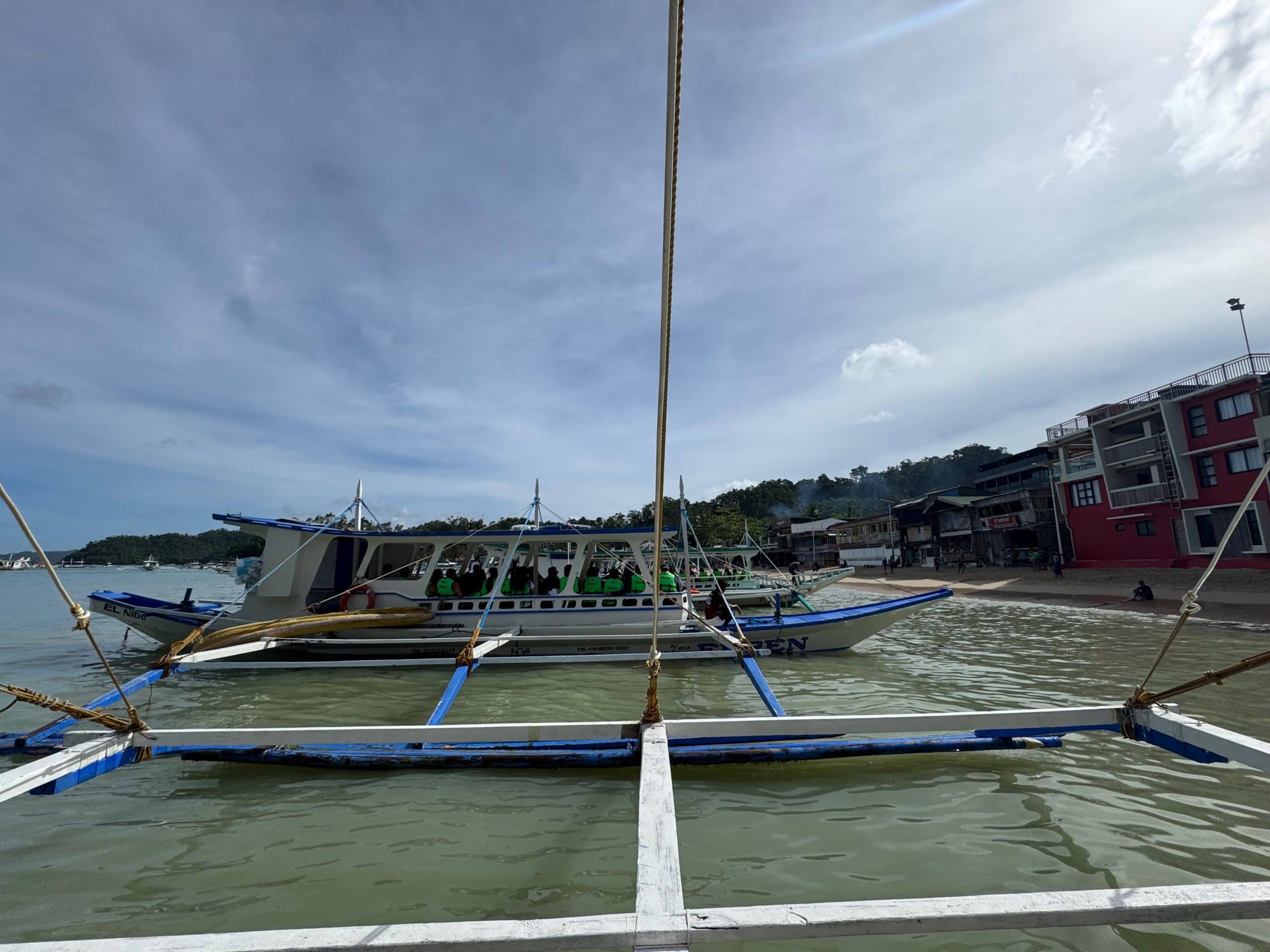 Traditional El Nido island hopping boat boarding near the shoreline with beachfront buildings and calm waters in Palawan Philippines