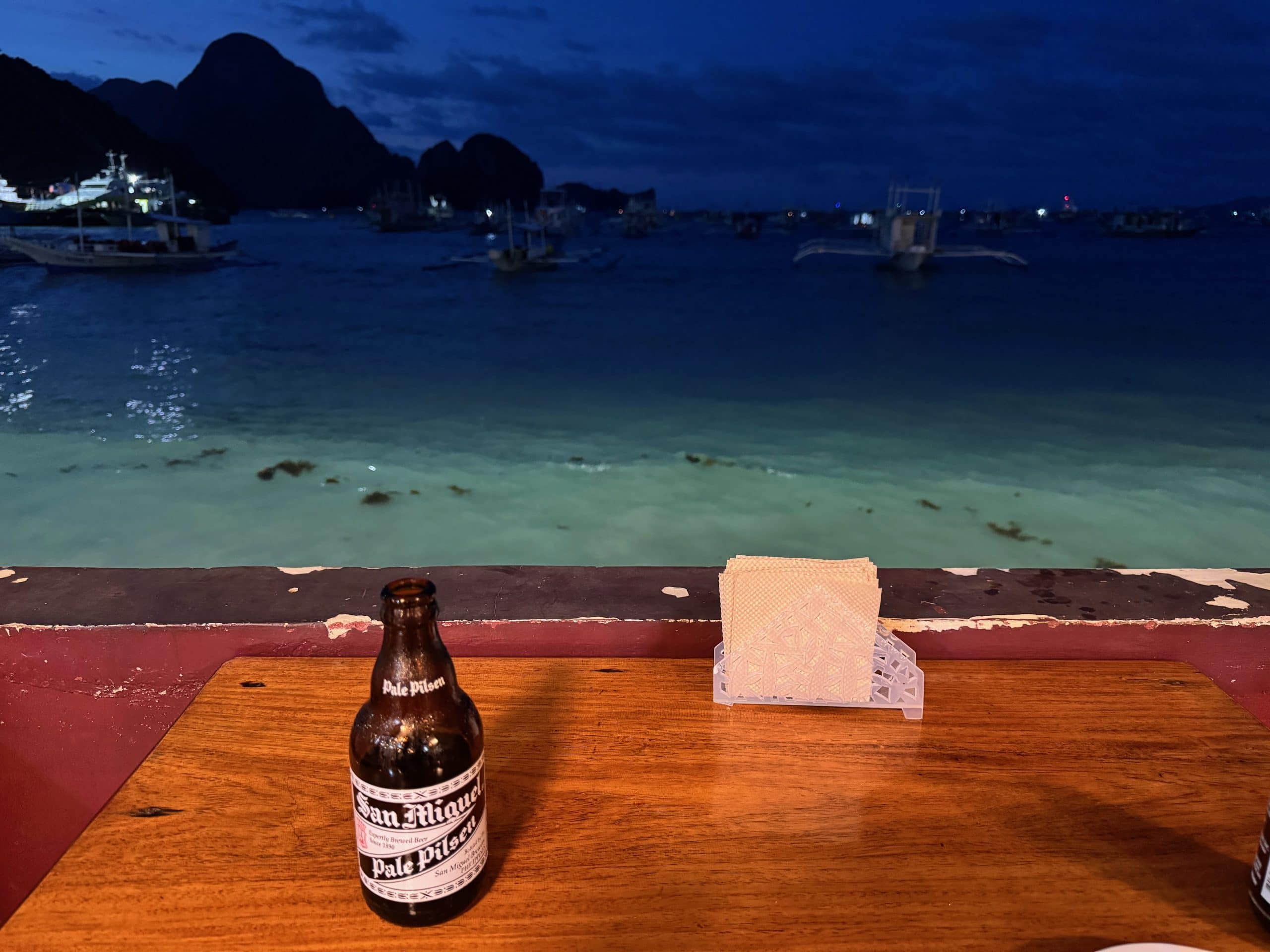 Cold San Miguel beer on a beachfront table in El Nido at night, overlooking calm turquoise water and island silhouettes in Palawan