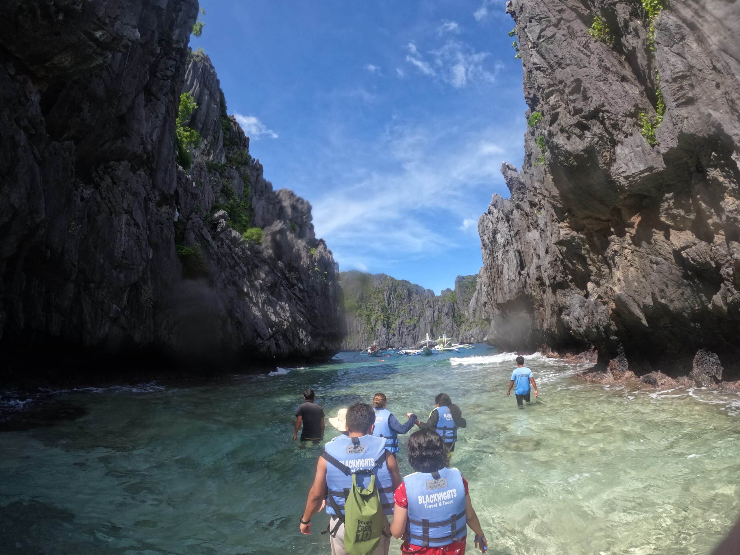Travelers walking through shallow turquoise water surrounded by towering limestone cliffs near Secret Lagoon during El Nido Tour A in Palawan Philippines.