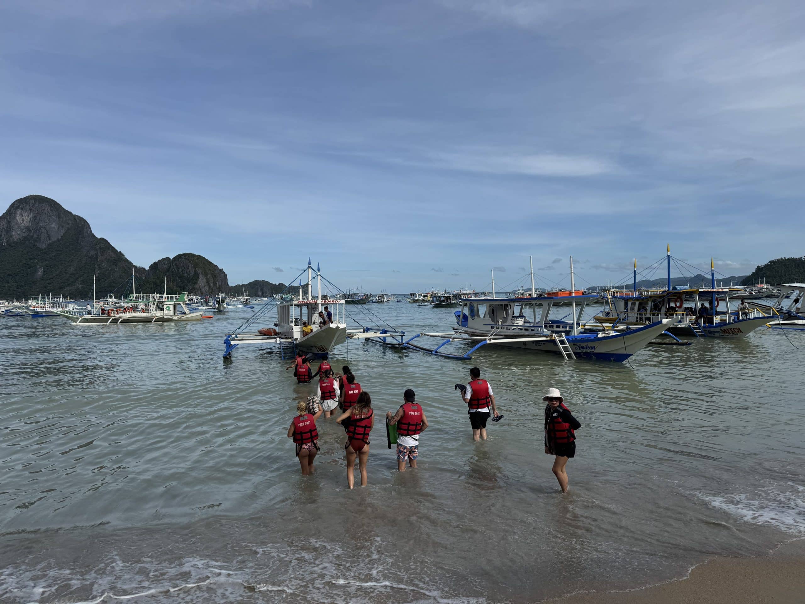Tourists walking through shallow water to board traditional island hopping boats in El Nido with limestone islands in the background in Palawan Philippines.