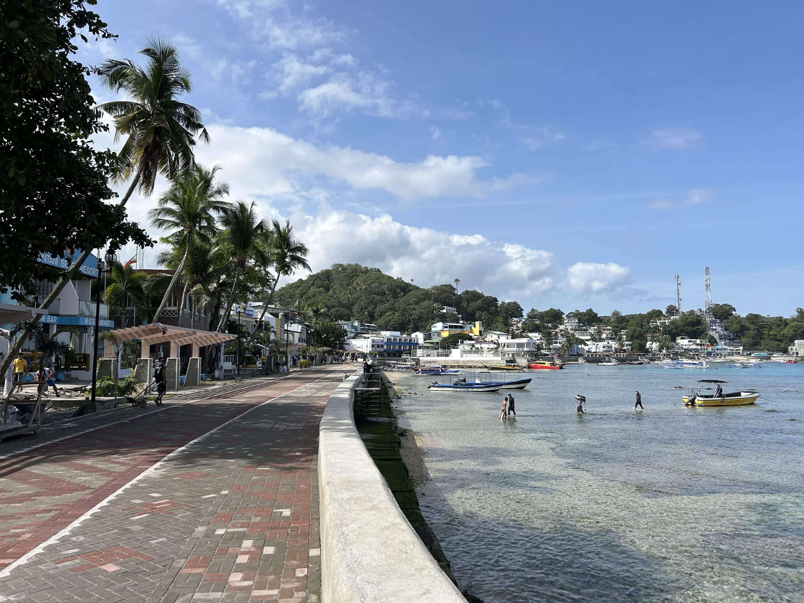 Waterfront promenade in Puerto Galera, Mindoro, with palm trees, local boats and clear water along the town’s coastline.