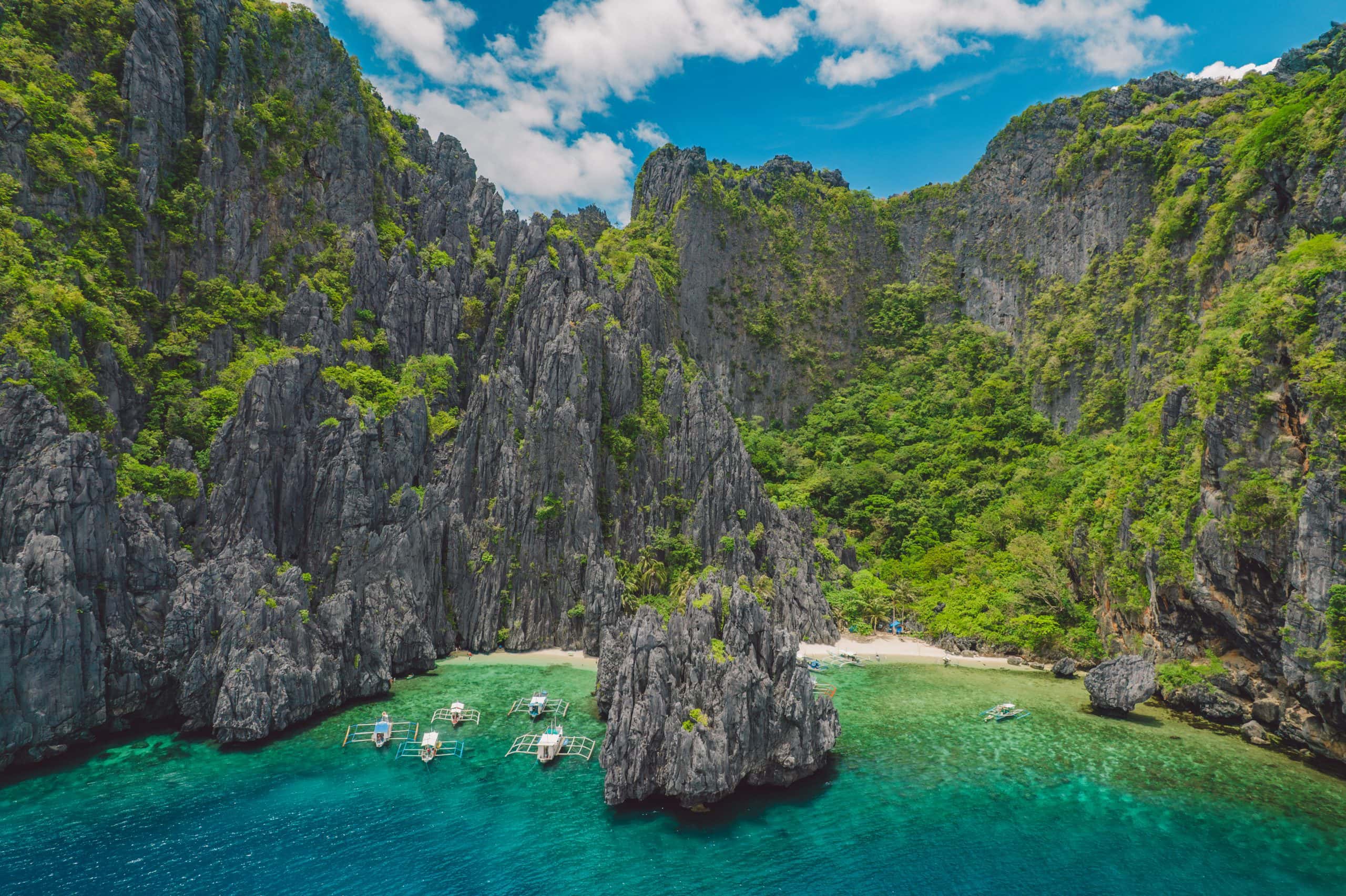 Aerial view of El Nido, Palawan, with turquoise water, limestone cliffs, tropical vegetation and traditional outrigger boats along the beach.